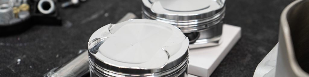 Two shiny new pistons resting on white bases on a workbench, surrounded by various engine parts and tools in a mechanic’s workshop.