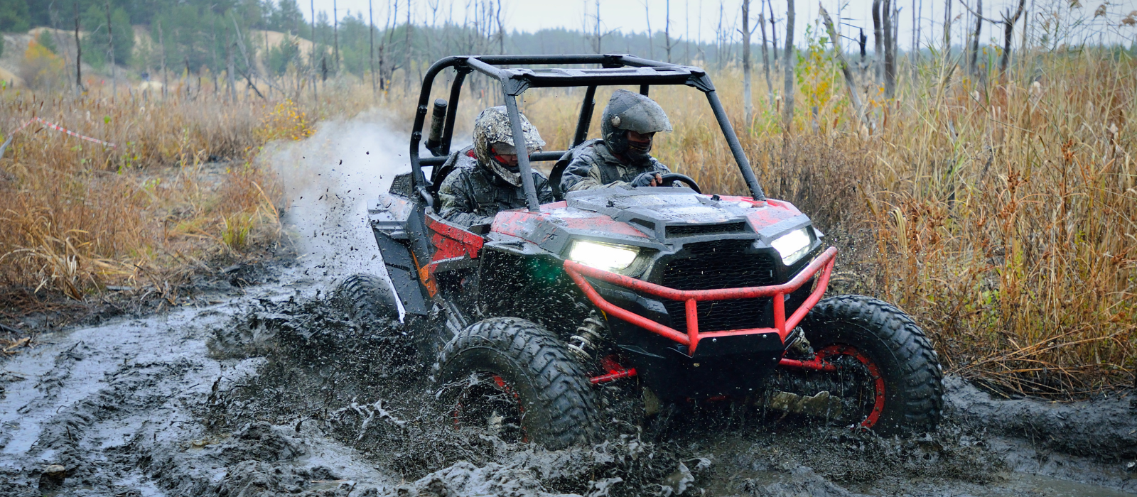 Polaris RZR powering through thick mud and water with two riders in full gear—built to handle brutal off-road terrain.